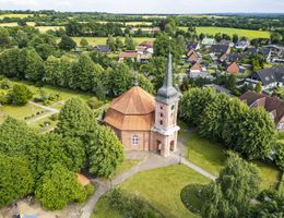 Luftaufnahme der Katharinenkirche in Großenaspe, umgeben von Bäumen und einem Friedhof, mit angrenzenden Wohnhäusern und Feldern im Hintergrund.