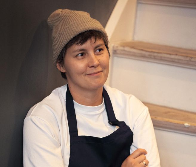 A person is sitting on a staircase wearing a cap and a work shirt. They look thoughtful and smile slightly.