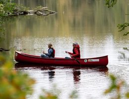 Zwei Personen paddeln in einem roten Kanu auf einem ruhigen Fluss, umgeben von grüner Vegetation.